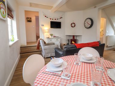 A dining table with bowls and glasses in front of a living room with chairs a fireplace and a wall clock at The Little White Cottage in Milton-Under-Wychwood
