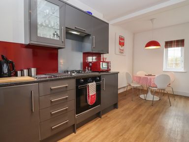 A kitchen with gray cabinets a red backsplash and a dining area with a round table and white chairs at The Little White Cottage in Milton-Under-Wychwood