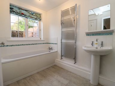 A bathroom with a bathtub, white pedestal sink, rectangular mirror, towel radiator, and window with patterned blind at The Little White Cottage in Milton-Under-Wychwood