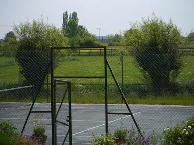 A tennis court behind a chain link fence with a gate open and greenery in the background at The Little White Cottage in Milton-Under-Wychwood