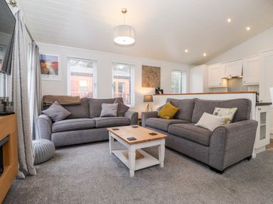 A living room with two gray sofas, a wooden coffee table, windows with blinds, and a view of an open kitchen at Bowness Bay Lodge Oakside 16 Fallbarrow Park near Bowness-On-Windermere