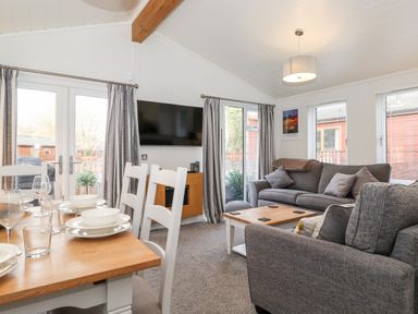 A living room with a dining table set with dishes and glasses a mounted TV grey sofas and coffee table at Bowness Bay Lodge Oakside 16 Fallbarrow Park near Bowness-On-Windermere