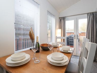 A dining table set with white plates and bowls glasses and a bottle near windows and glass doors at Bowness Bay Lodge Oakside 16 Fallbarrow Park near Bowness-On-Windermere