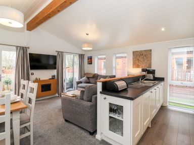 An open living room and kitchen area with gray sofas a wall-mounted TV dining table and white kitchen cabinets at Bowness Bay Lodge Oakside 16 Fallbarrow Park near Bowness-On-Windermere