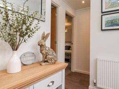 A hallway with a wooden table holding a vase with branches, a wooden rabbit sculpture, and a decorative sign with a bedroom visible through an open door at Bowness Bay Lodge Oakside 16 Fallbarrow Park near Bowness-On-Windermere