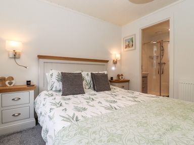 A bedroom with a double bed with leaf patterned bedding two bedside tables wall lamps and an en suite shower at Bowness Bay Lodge Oakside 16 Fallbarrow Park near Bowness-On-Windermere