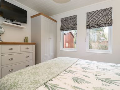 A bedroom with a bed covered in patterned bedding two windows with patterned blinds a white wardrobe and chest of drawers with a television mounted above at Bowness Bay Lodge Oakside 16 Fallbarrow Park near Bowness-On-Windermere