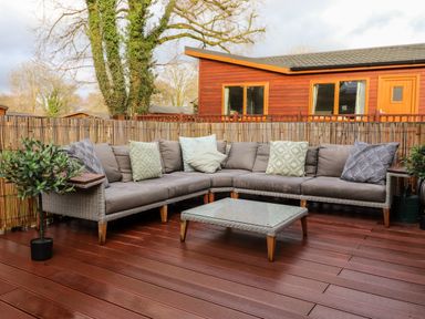 An outdoor seating area with a grey sectional sofa cushions glass coffee table and two potted plants on wooden decking at Bowness Bay Lodge Oakside 16 Fallbarrow Park near Bowness-On-Windermere