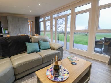 A living room with a gray leather sofa and coffee table with a bottle and glasses by large windows at Holmside Audley Brow near Market Drayton