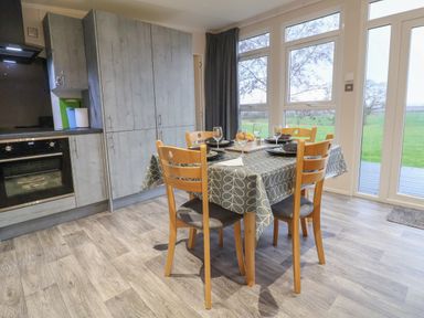 A dining area with a table covered with a patterned cloth set with plates and glasses and wooden chairs next to a kitchen with grey cabinets and a large window showing a green field at Holmside Audley Brow near Market Drayton