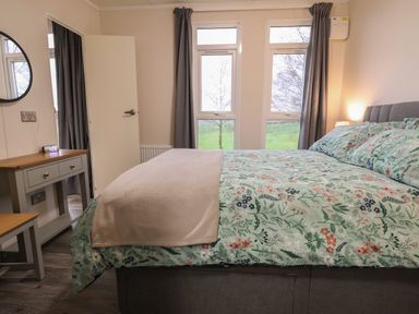 A bedroom with a floral bedspread a bedside lamp a small table with a mirror and alarm clock and two windows with curtains at Holmside Audley Brow near Market Drayton