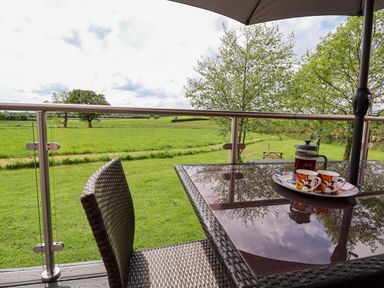 An outdoor balcony with a glass table, two cups on a tray, a coffee press, a wicker chair, and a green field with trees in the background at Holmside in Audley Brow near Market Drayton