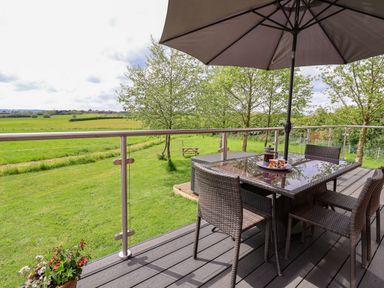 An outdoor deck with a glass table and four wicker chairs under an umbrella overlooking a grassy field with trees at Holmside Audley Brow near Market Drayton