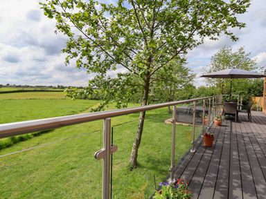 A wooden deck with metal and glass railing overlooking a green field with trees at Holmside in Audley Brow near Market Drayton