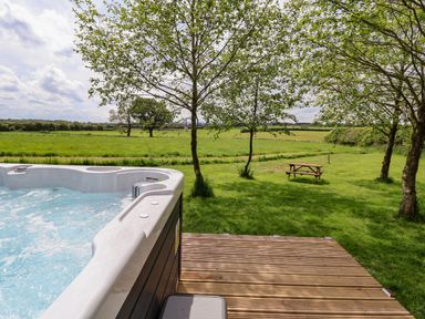 An outdoor hot tub next to a wooden deck with trees and a picnic bench on a grassy field at Holmside in Audley Brow near Market Drayton