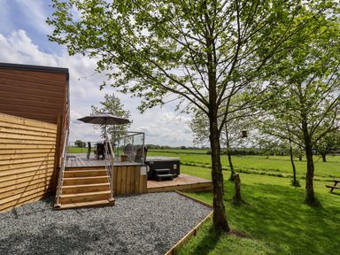 A wooden deck with stairs, an umbrella, table and chairs, a hot tub next to a grassy area with trees at Holmside in Audley Brow near Market Drayton