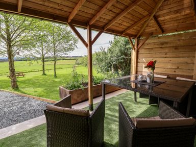 An outdoor covered seating area with a glass table and wicker chairs overlooking a grassy field at Holmside in Audley Brow near Market Drayton