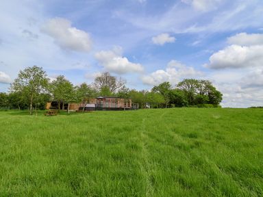 A wooden cabin with a deck and picnic table in a large grassy field with trees under a blue sky at Holmside Audley Brow near Market Drayton