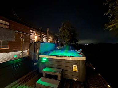 An outdoor hot tub with green lights on a wooden deck at night near a building with lit windows at Holmside in Audley Brow near Market Drayton