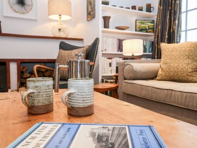A living room with a wooden table holding two mugs a coffee press and a book the room has a sofa with cushions a chair a lamp and built in bookshelves at Beta Cottage in Walberswick