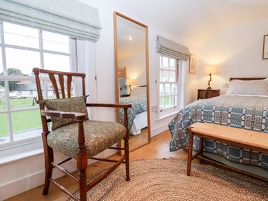 A bedroom with a wooden chair near windows a full-length mirror a bed with patterned bedding and a bench at Beta Cottage in Walberswick