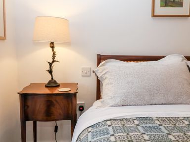 A wooden bedside table with a decorative lamp and a bed with patterned pillows and a blanket at Beta Cottage in Walberswick