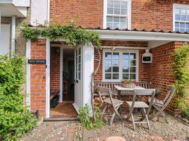 An outdoor seating area with a wooden table and chairs outside a brick cottage with an open door and climbing plants at Beta Cottage in Walberswick