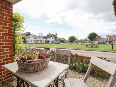 A small outdoor seating area with a table and two chairs overlooking a grassy park with houses and parked cars at Beta Cottage in Walberswick