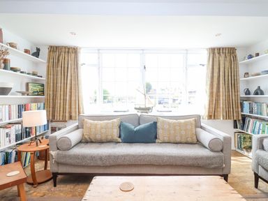A living room with a gray sofa and patterned cushions in front of a large window with yellow curtains and bookshelves on each side at Beta Cottage in Walberswick