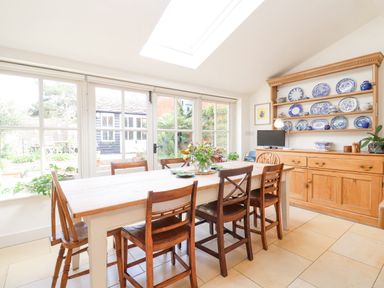 A dining room with wooden table and chairs next to large windows and a wooden cabinet with blue and white plates at Beta Cottage in Walberswick