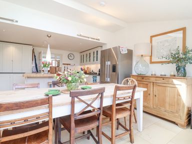 A dining area with a wooden table and chairs a sideboard with a lamp flowers and a refrigerator in the background at Beta Cottage in Walberswick