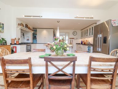 A kitchen with a wooden dining table and chairs a flower vase on the table white cabinets a stainless steel refrigerator and a clock on the wall at Beta Cottage in Walberswick