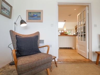 A patterned armchair with a black cushion and a floor lamp next to it in a sitting room with a view into a kitchen with a white island at Beta Cottage in Walberswick