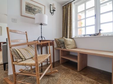 A sitting area with a wooden rocking chair with striped cushion a wooden bench with pillows a small wooden table with a table lamp and a window with patterned curtains at Beta Cottage in Walberswick