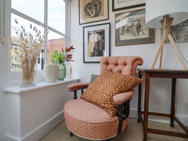 A pink upholstered armchair with throw pillows next to a wooden table with a lamp and a window sill with plants and dried flowers at Beta Cottage in Walberswick