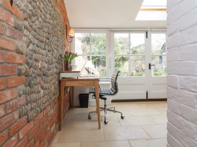 A small workspace with a wooden desk and black chair next to a pebble and brick wall with a windowed door to a garden at Beta Cottage in Walberswick