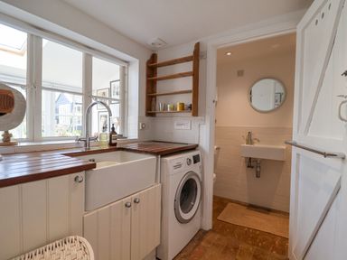 A utility room with a sink washing machine wooden shelves and a doorway leading to a bathroom with a sink and a round mirror at Beta Cottage in Walberswick