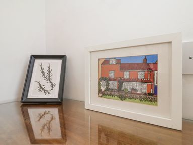 Two framed pictures on a wooden surface one with a plant drawing and the other with a house illustration at Beta Cottage in Walberswick