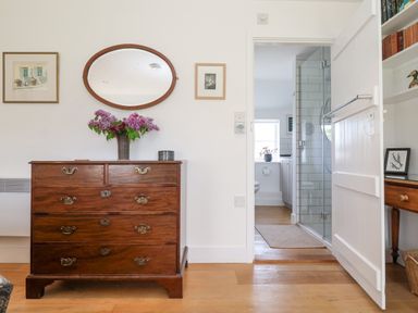 A wooden dresser with flowers in a vase a mirror above and a bathroom visible through an open door at Beta Cottage in Walberswick