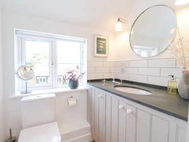 A bathroom with a toilet below a window a round mirror above a sink and countertop at Beta Cottage in Walberswick