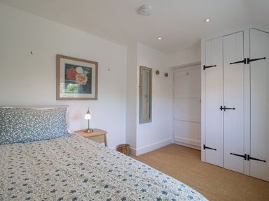 A bedroom with a patterned bedspread a framed floral picture a bedside table with a lamp a mirror and white storage cupboards at Beta Cottage in Walberswick