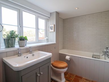 A bathroom with a sink vanity and plants on the windowsill a toilet with a wooden seat a bathtub with tiled walls at Beta Cottage in Walberswick