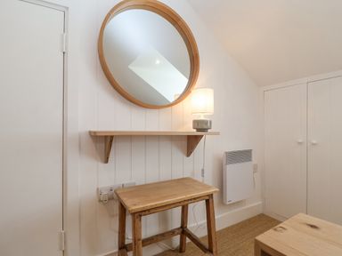 A wooden stool below a wooden shelf with a lamp and round wooden framed mirror on the wall in a white room at Beta Cottage in Walberswick