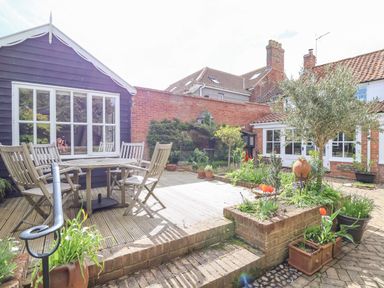 A garden patio with wooden table and chairs surrounded by plants and trees at Beta Cottage in Walberswick