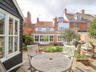 A backyard patio with a wooden table and chairs surrounded by plants and a brick house at Beta Cottage in Walberswick