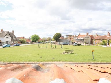 A grassy park area with swings and a bench surrounded by houses and parked cars at Beta Cottage in Walberswick