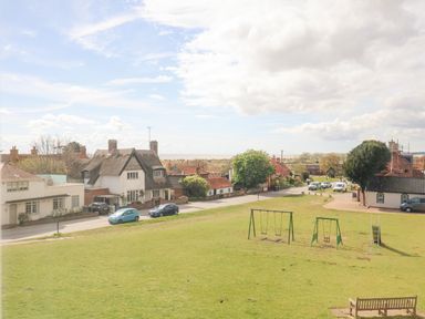 A grassy park area with swings and a slide near a road with houses and parked cars at Beta Cottage in Walberswick