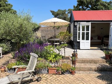 A garden patio with wooden chairs and table under an umbrella near a small black building with double white doors at Beta Cottage in Walberswick