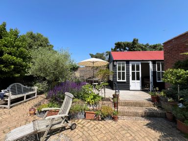 A garden with wooden chairs and a table with an umbrella near a black shed with a red roof at Beta Cottage in Walberswick
