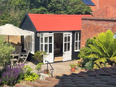 A small garden shed with red roof and black walls with white doors and windows surrounded by plants and outdoor furniture at Beta Cottage in Walberswick